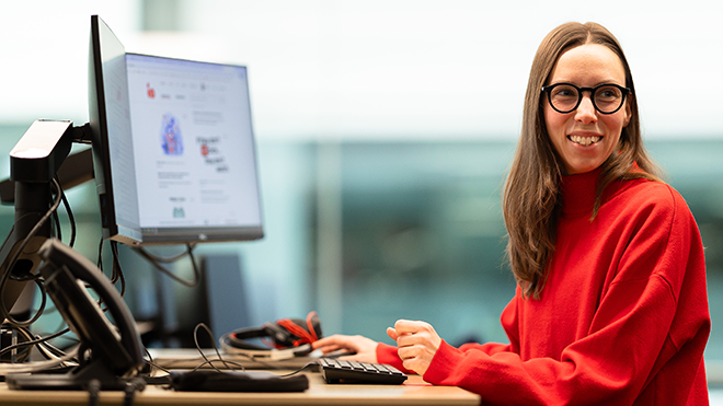 Une femme aux longs cheveux bruns et aux lunettes rondes noires sourit en détournant le regard de ses écrans d’ordinateur. Elle porte un pull rouge vif et se tient debout à un bureau à hauteur ajustable dans un espace de bureau moderne et bien éclairé.