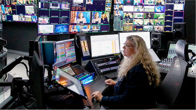 Une femme aux longs cheveux blonds ondulés est assise, concentrée, dans une salle de contrôle, entourée de plusieurs écrans affichant de multiples vidéos et informations.