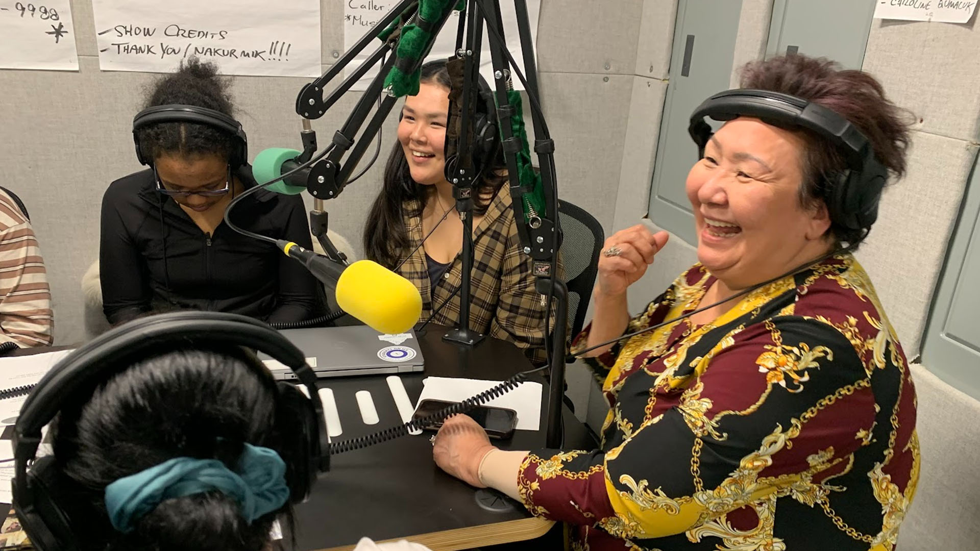 Deux étudiantes et première réalisatrice de CBC North Salome Avva sont assises autour d’une table souriantes, devant des microphones et portant des écouteurs.