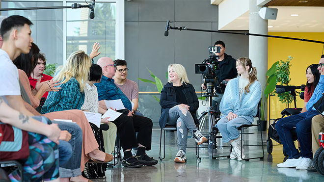 Un groupe de personnes autistes et neurodivergentes en pleine discussion et assis en cercle dans un atrium lumineux et moderne. En arrière-plan, un caméraman filme le groupe avec une caméra professionnelle.
