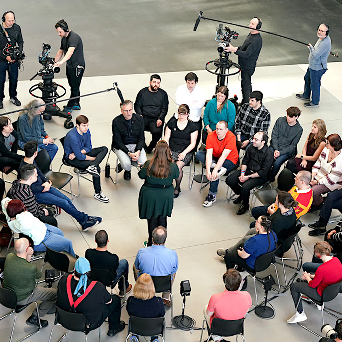 Une vue en plongée du  tournage d’une émission qui montre un groupe de personnes autistes et neurodivergentes assises en cercle dans une pièce spacieuse et lumineuse. Une femme au centre du cercle est interviewée par le groupe, pendant que des caméramans professionnels filment la discussion.