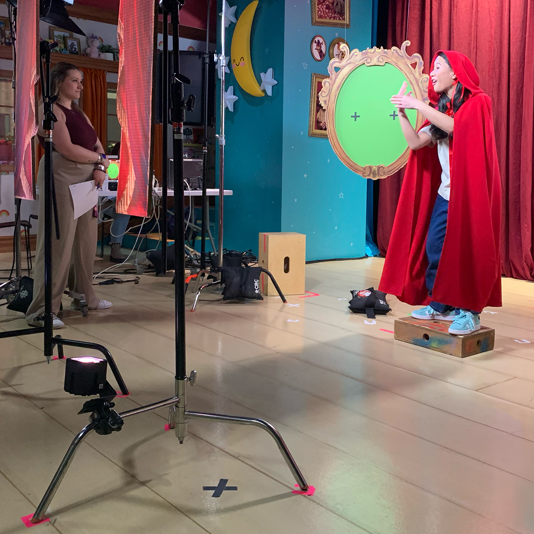 Une comédienne dans un studio de télévision déguisée en Petit Chaperon rouge fait des gestes vers un cadre de miroir orné avec un fond vert.