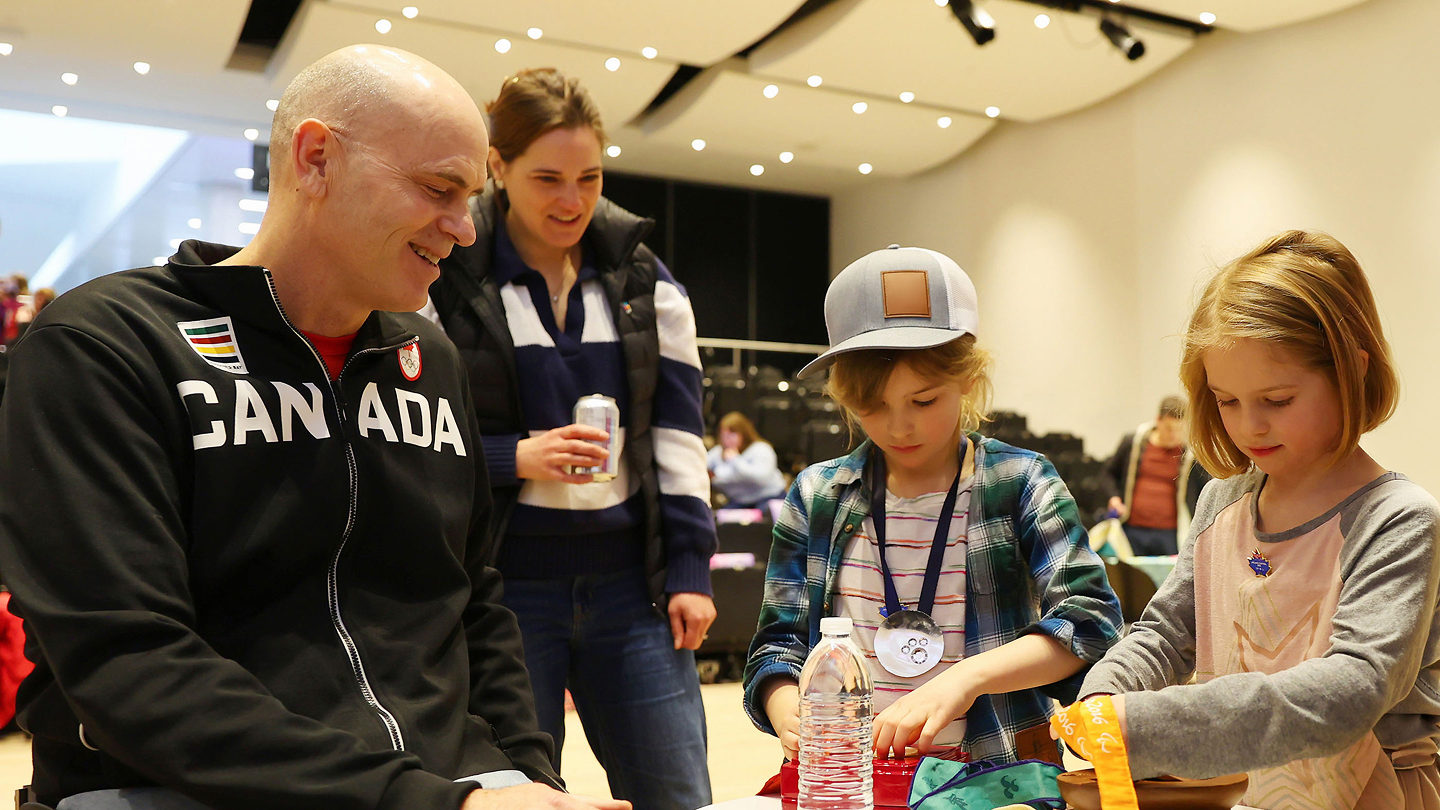 L'athlète paralympique Paul Tingley, assis en fauteuil roulant et portant un chandail à capuchon noir d'Équipe Canada, en train de confectionner des médailles avec deux jeunes enfants.