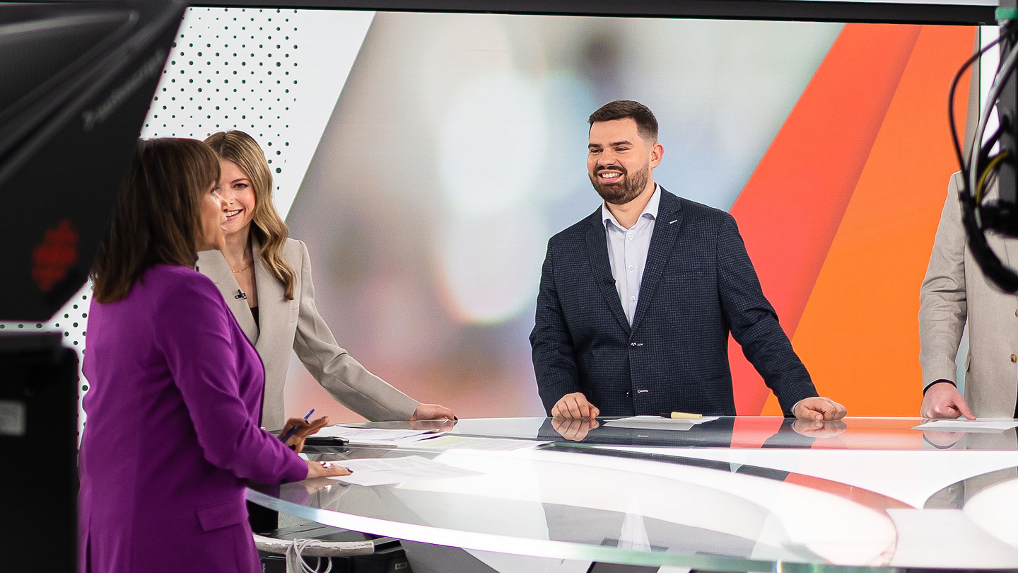Une vue des coulisses d'un studio de nouvelles télévisées très éclairé. Quatre présentateurs se tiennent debout, sourient et discutent derrière un bureau moderne en verre incurvé.