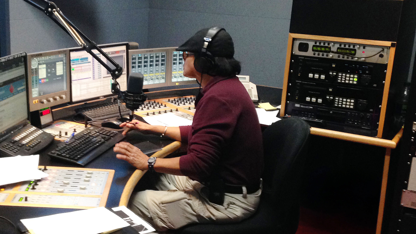 Un homme portant une casquette et un casque d'écoute regarde vers les moniteurs et utilise une grande console de mixage audio à plusieurs écrans. Une télévision fixée au mur au-dessus de lui montre un skieur alpin en pleine action.